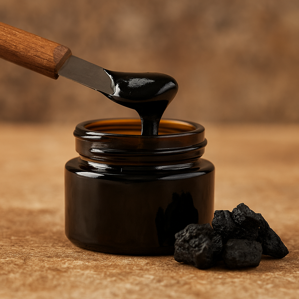 A high-resolution photograph displays a small, round amber glass jar filled with thick black shilajit resin. A wooden-handled spatula lifts the glossy resin from the jar, with solid pieces of shilajit resting nearby on a warm, earthy-toned surface.