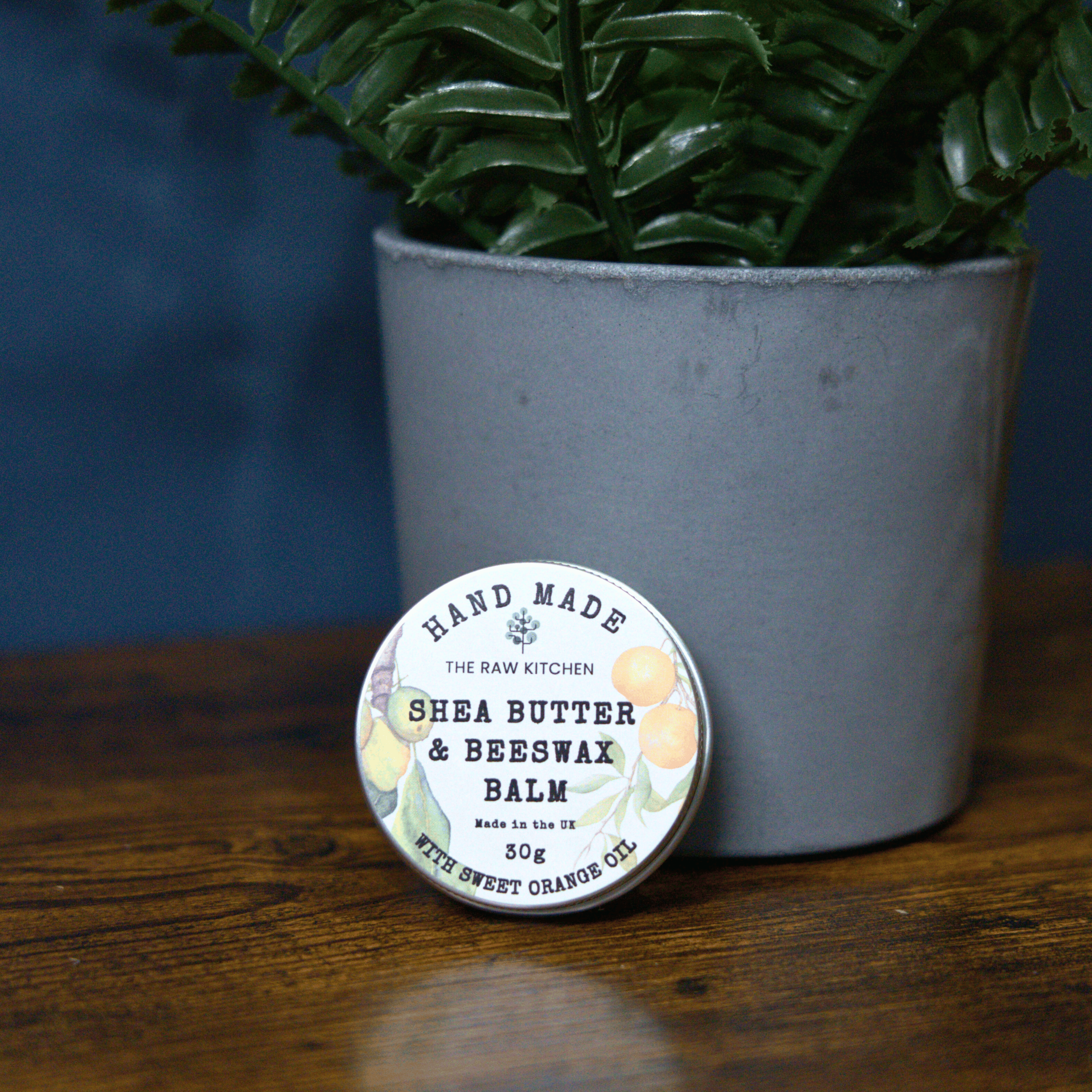 The handmade Shea Butter & Beeswax Balm tin shown on a wooden table next to a plant - The Raw Kitchen UK.