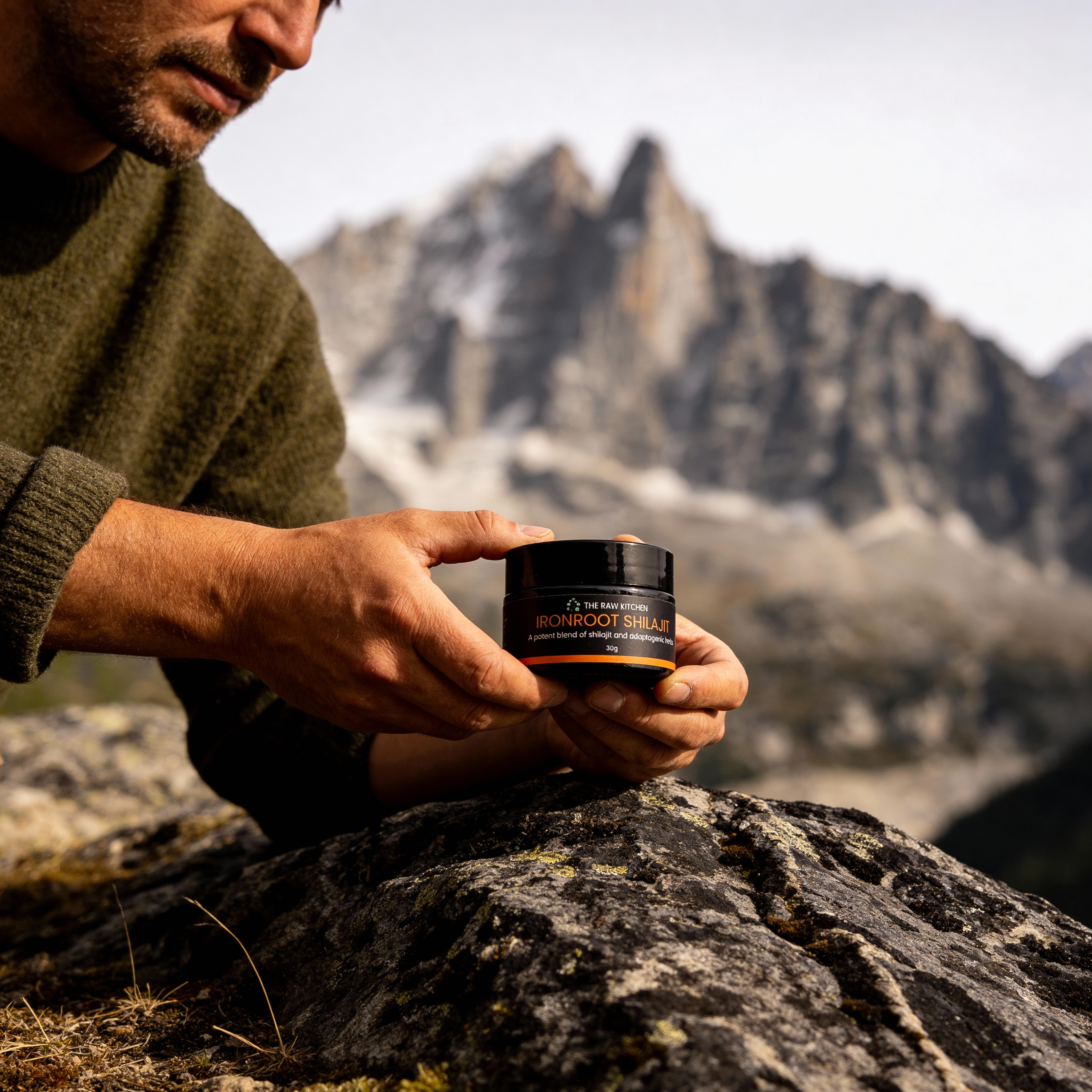 a man holding a jar of shilajit while leaning on a stone in a mountainous area.