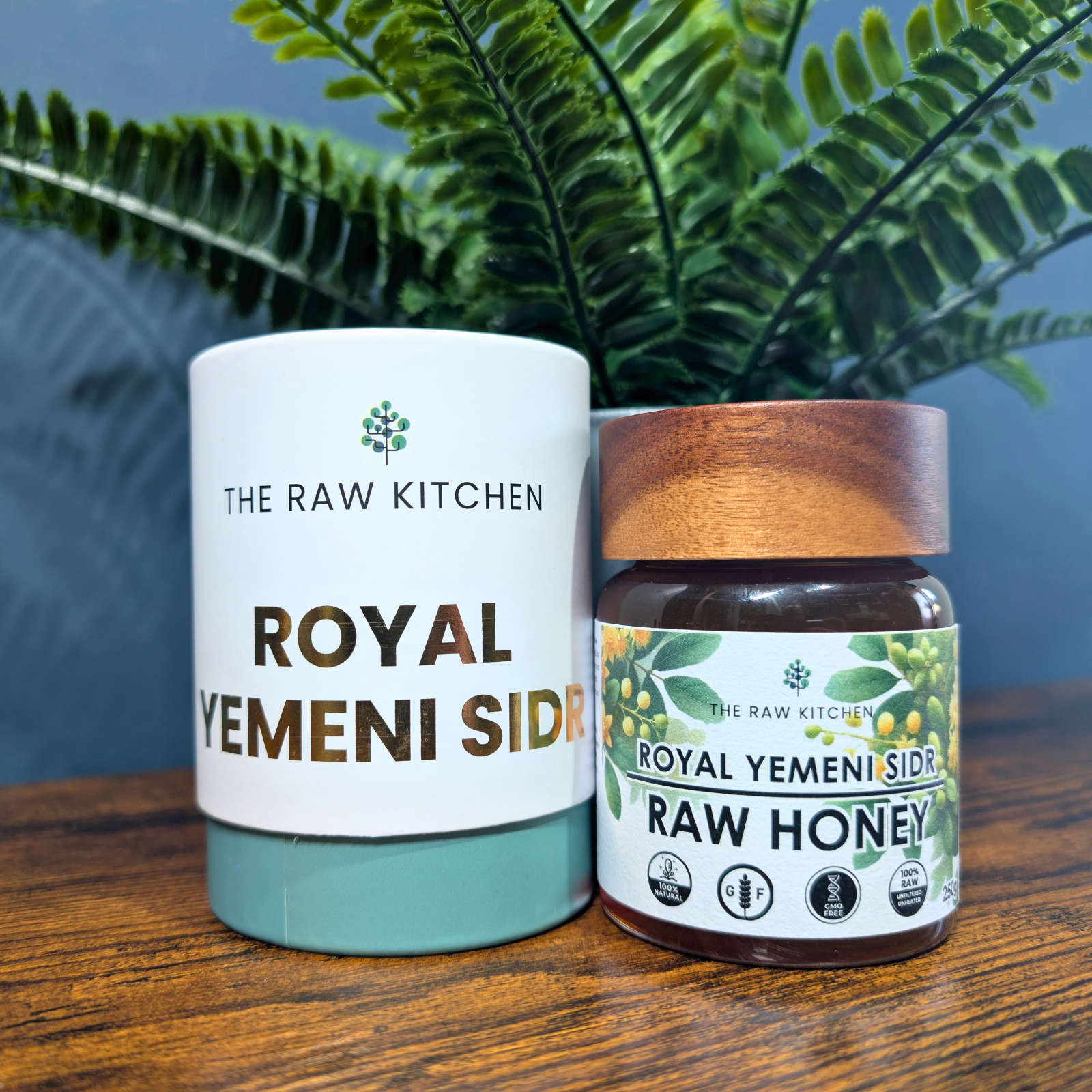 A 250g glass jar of Royal Yemeni Sidr Raw Honey with a Wooden lid and branded label next to a white branded box. A plant sits in the background - The Raw Kitchen UK.