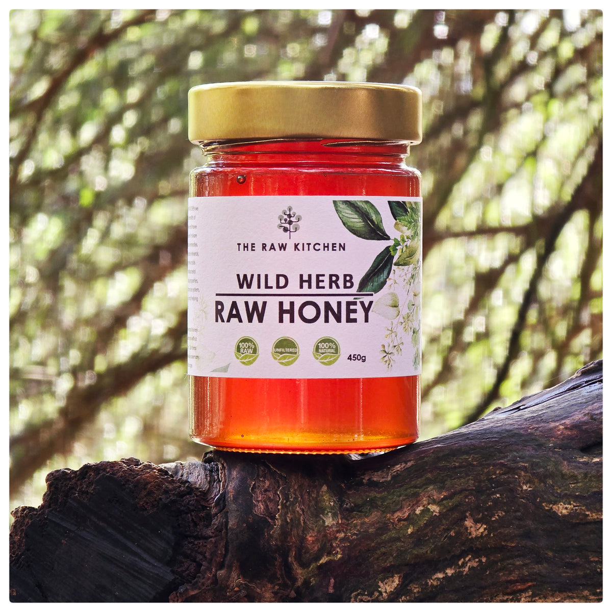 A jar of Wild Herb Raw Honey from The Raw Kitchen UK, resting on a rustic log in a natural, outdoor setting.