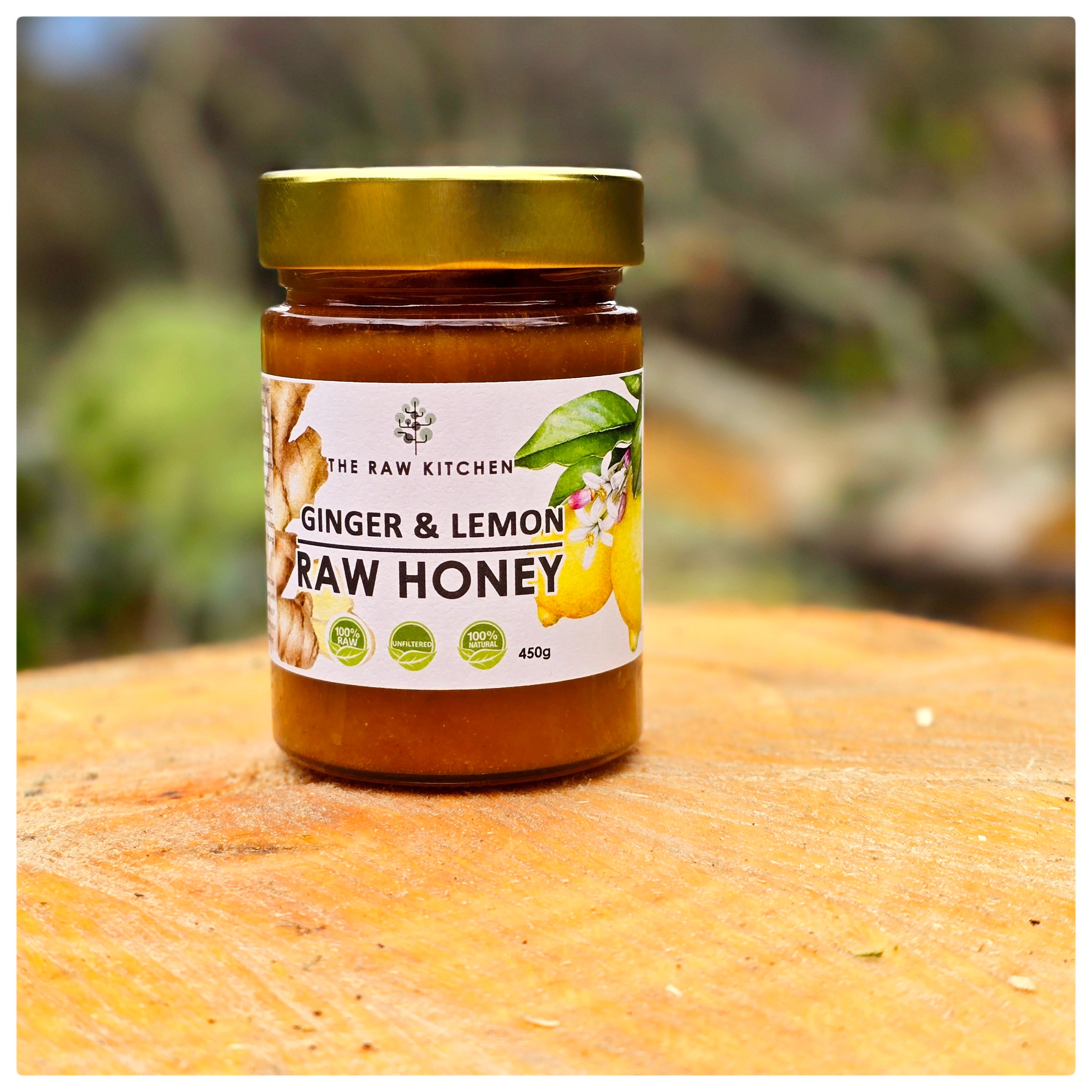 A 450g glass jar of Ginger & Lemon Raw Honey with a gold lid and branded label palced on a piece of wood in a garden - The Raw Kitchen UK.