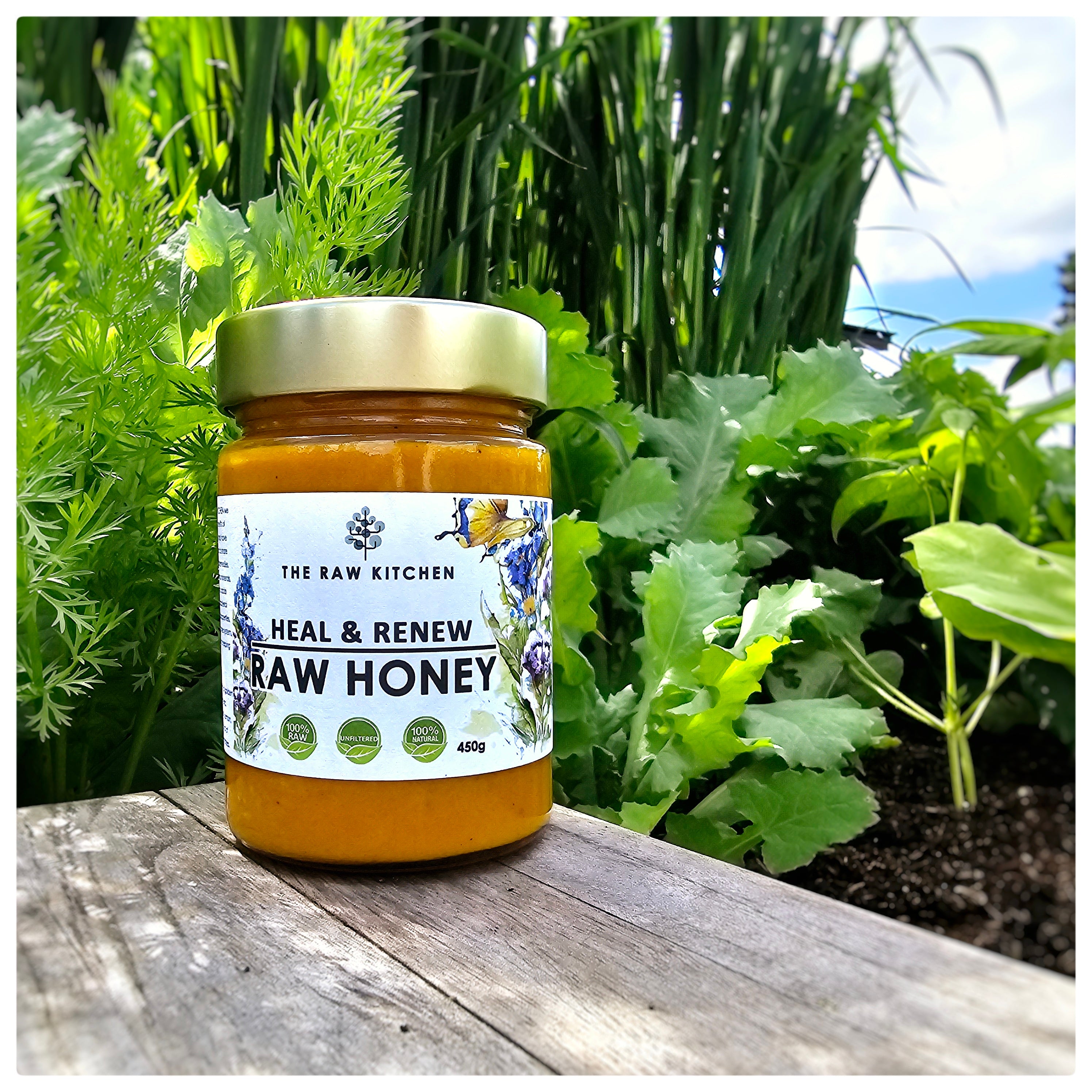 A 450g glass jar of Heal & Renew Raw Honey with a gold lid and branded label placed on a wooden board behind some plants - The Raw Kitchen UK.