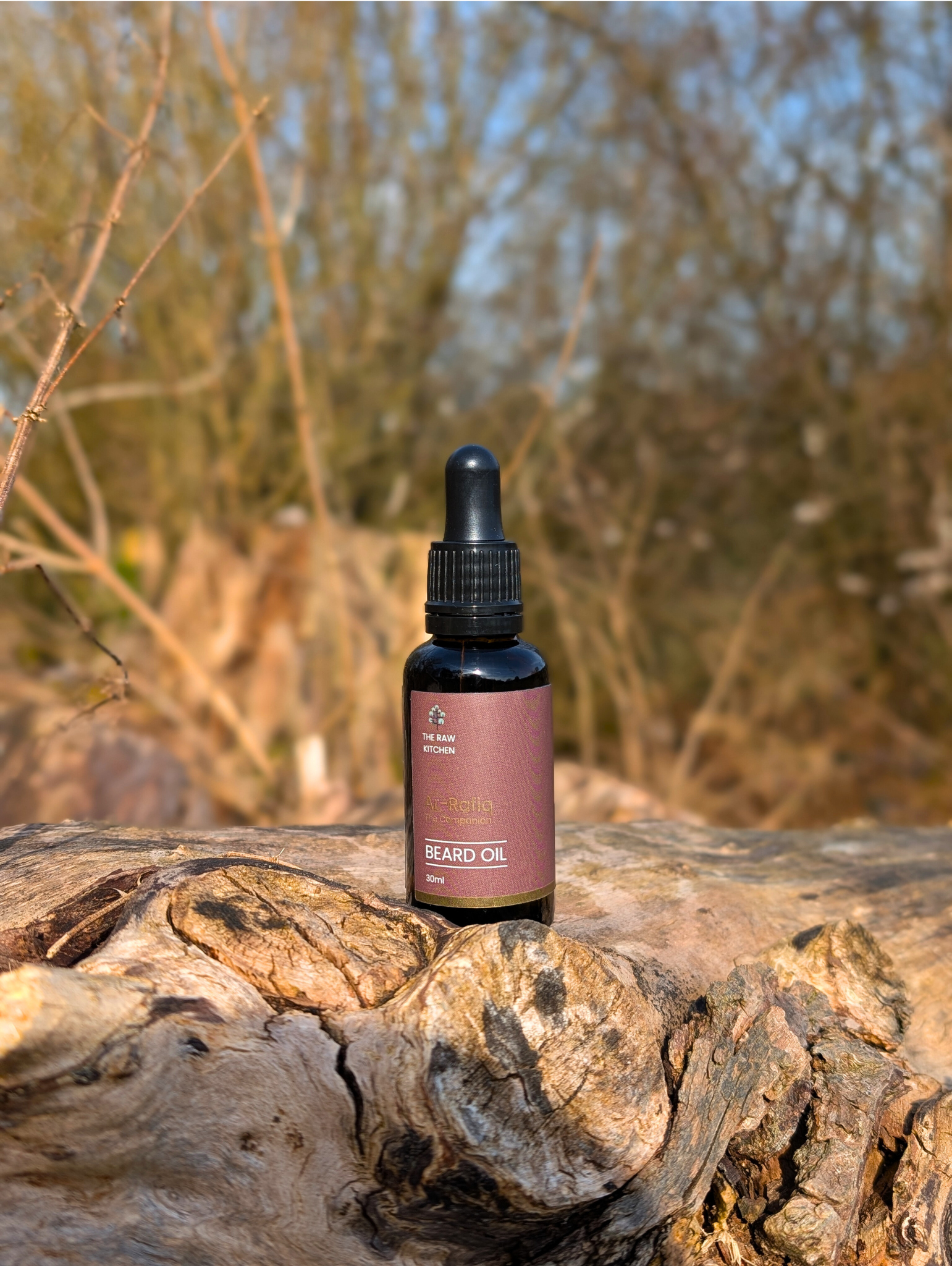A bottle of Ar-Rafiq "The Companion" Beard Oil resting on a rustic log in a natural outdoor setting - The Raw Kitchen UK.