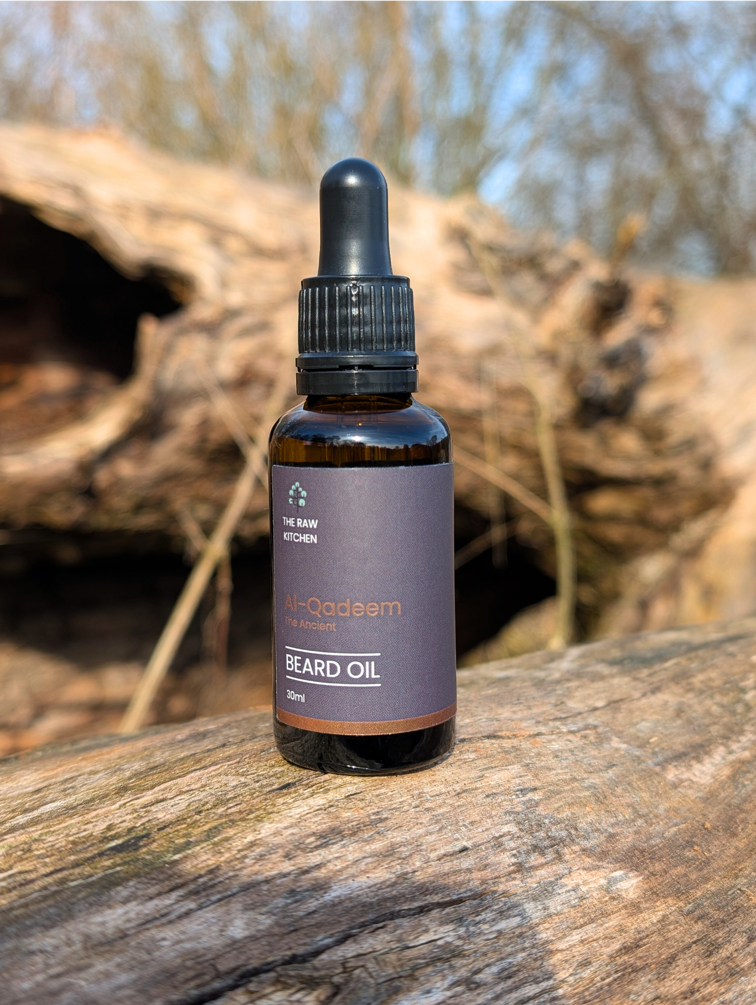 A bottle of Al-Qadeem "The Ancient" Beard Oil resting on a rustic log in a natural setting - The Raw Kitchen UK.