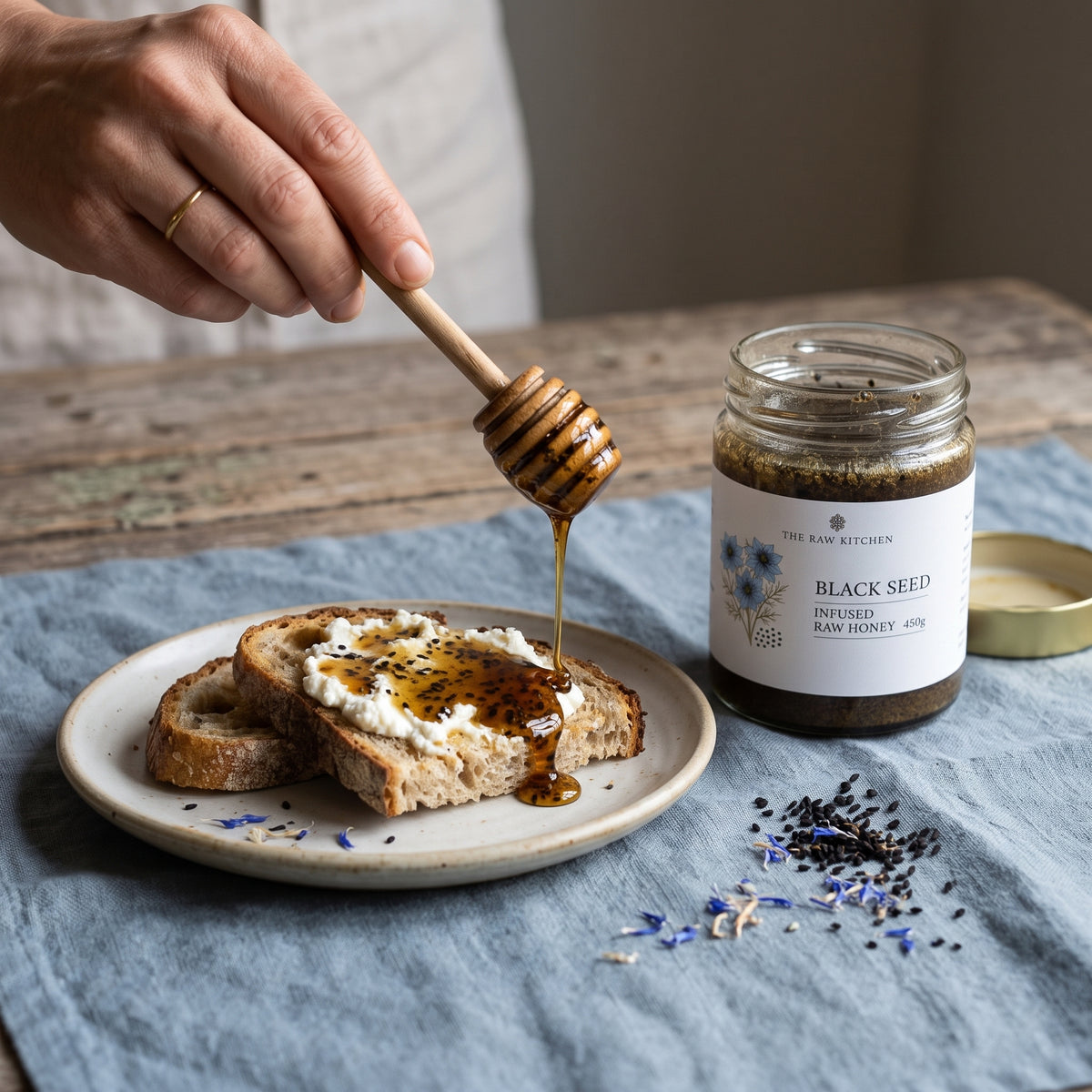 A hand drizzles honey from a wooden dipper onto toast on a plate, with a jar of The Raw Kitchen Black Seed Infused Raw Honey nearby on a blue cloth.