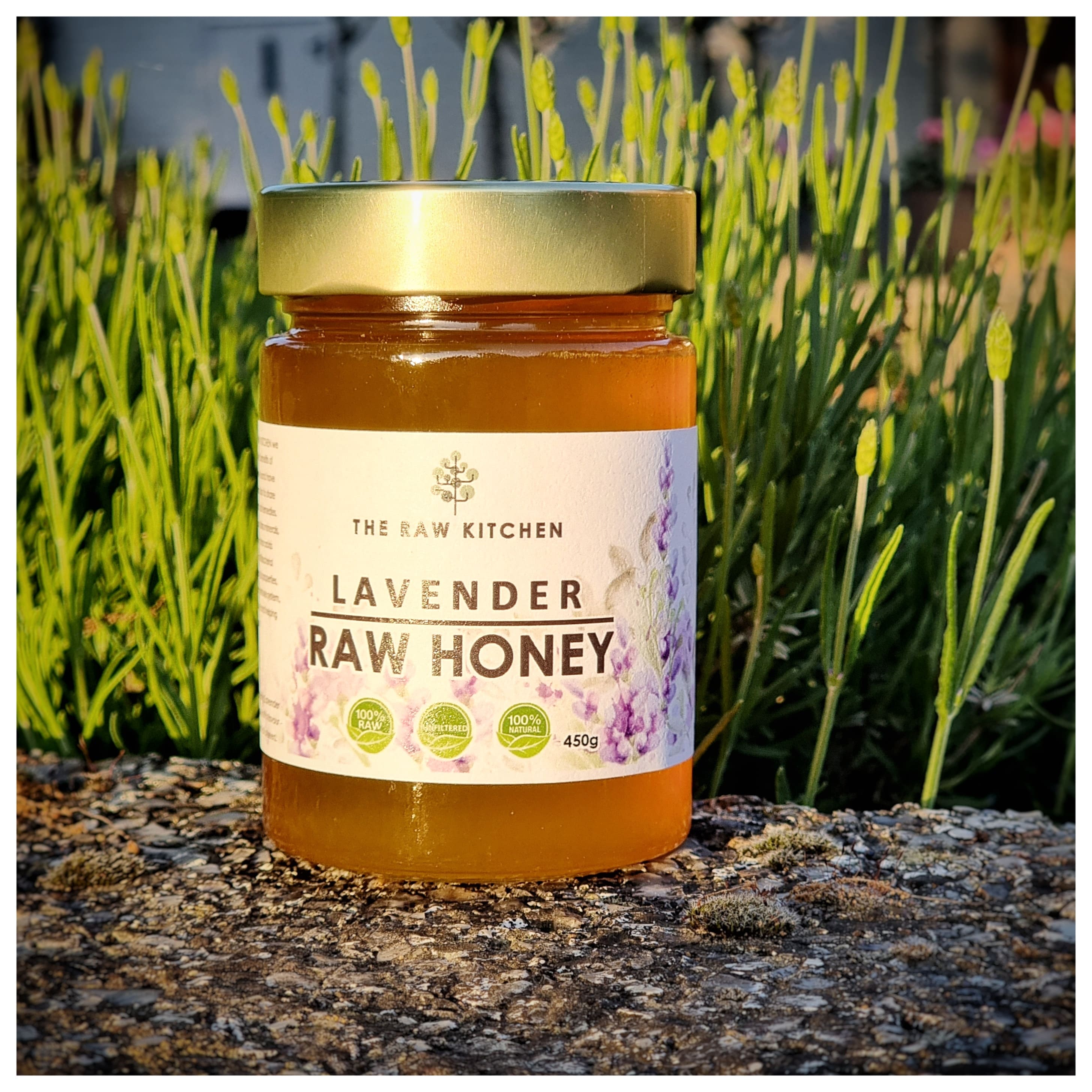 A 450g glass jar of Lavender Raw Honey with a gold lid and branded label placed on a stone infront of some herbs - The Raw Kitchen UK.