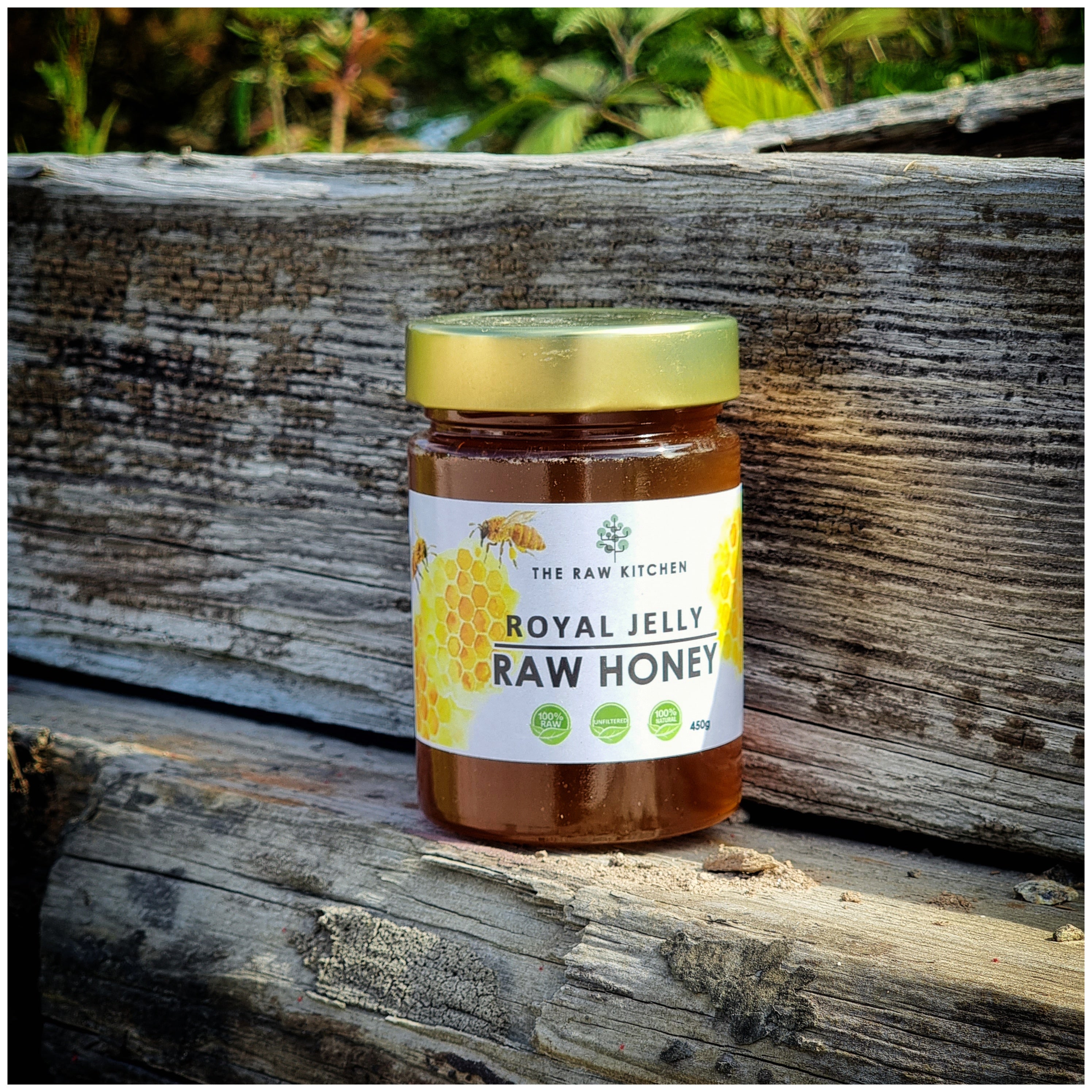 A 450g glass jar of Royal Jelly Infused Raw Honey with a gold lid and branded label placed on a wooden stump in a forest - The Raw Kitchen UK.