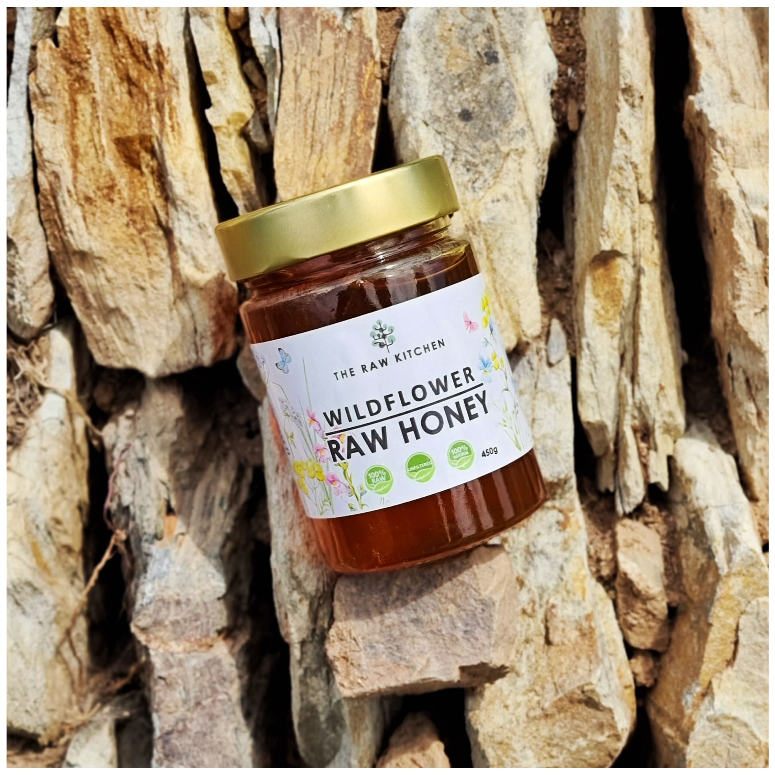 A 450g glass jar of Wildflower Raw Honey with a gold lid and branded label placed on some stones - The Raw Kitchen UK.