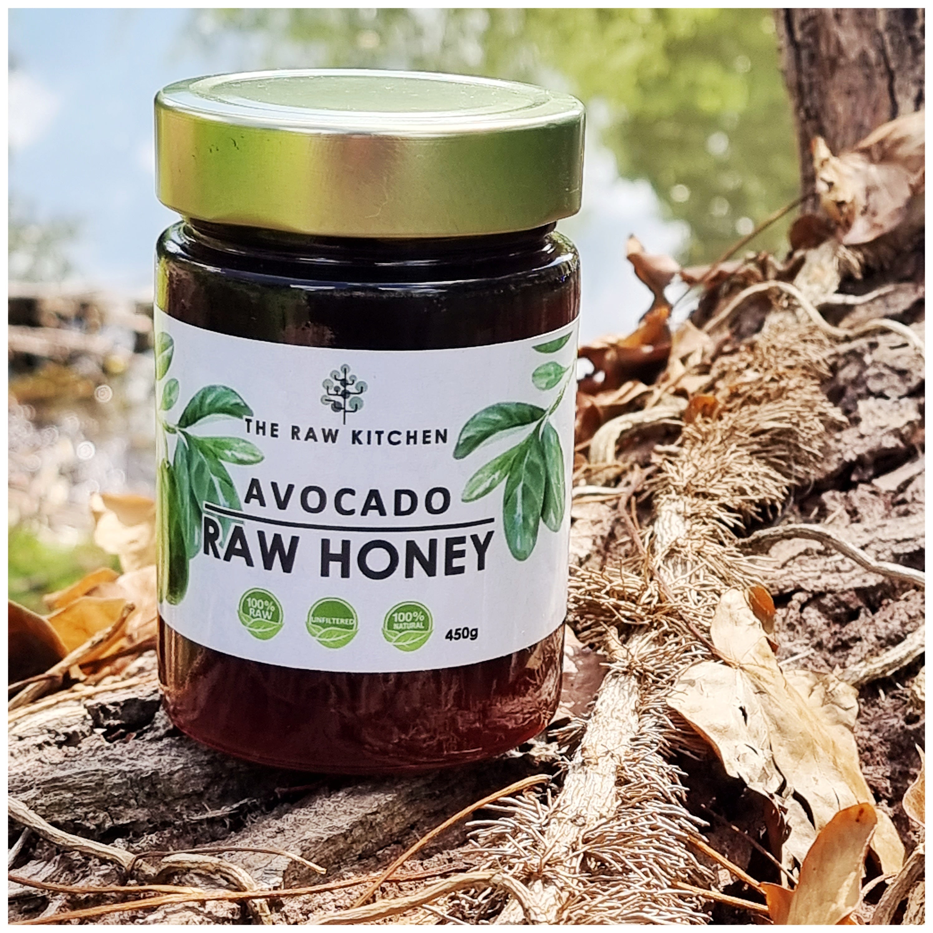 A 450g glass jar of Avocado Raw Honey with a gold lid and branded label placed on a tree stump in a forest- The Raw Kitchen UK.