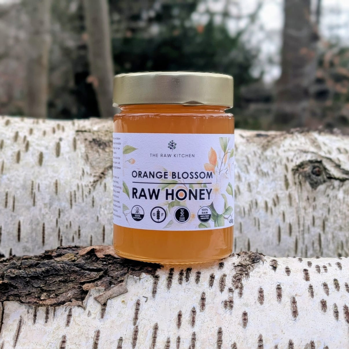A 450g glass jar of Orange Blossom Raw Honey with a gold lid and branded label placed on a tree stump in a forest - The Raw Kitchen UK.