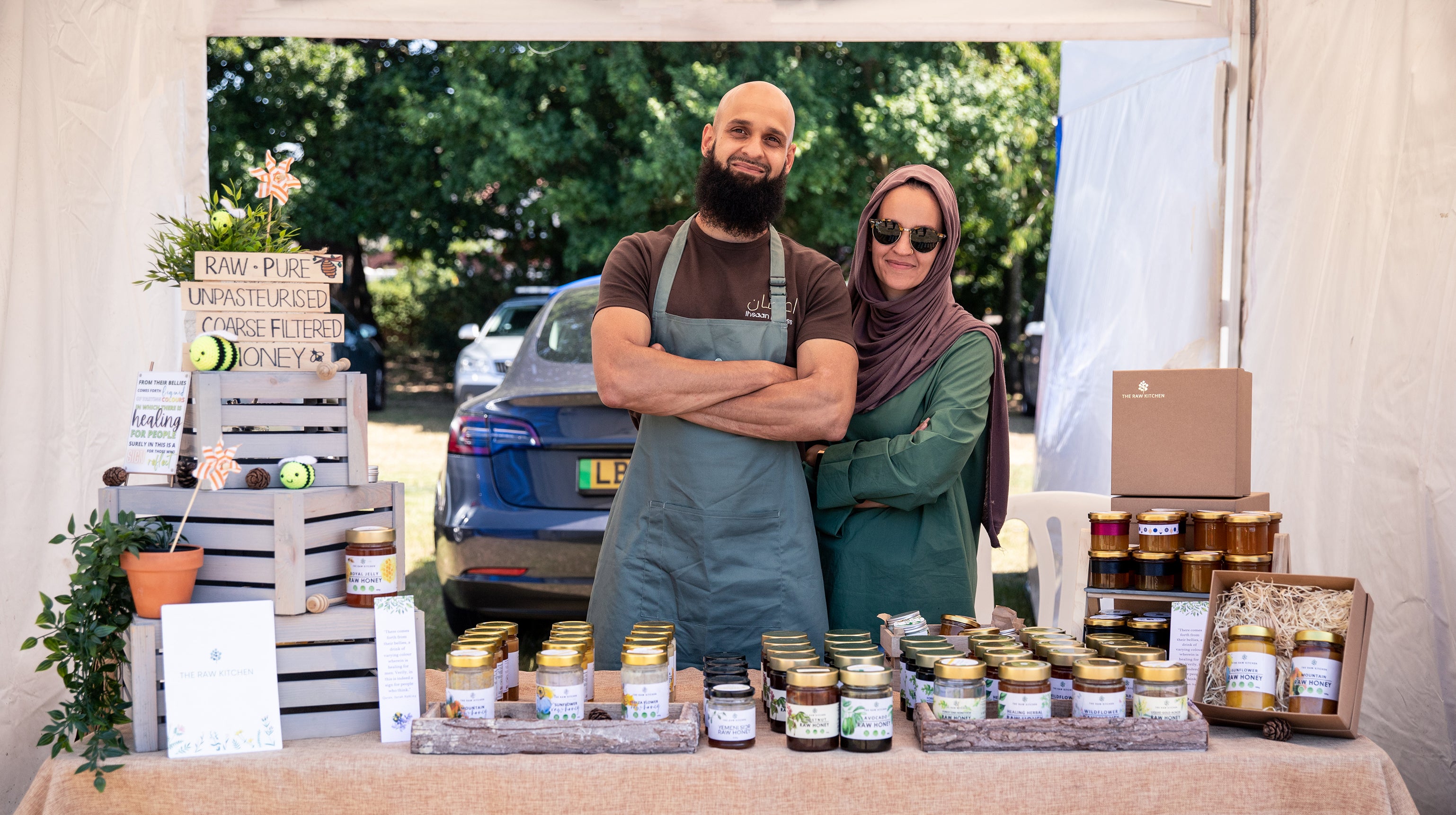 The founder of The Raw Kitchen and his partner smiling behind their beautifully arranged market stall, ready to serve customers jars of raw honey and other natural products.