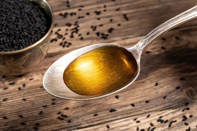 A close-up of pure black seed oil on a silver spoon, showing its golden colour, with raw black seeds in the background - The Raw Kitchen UK.