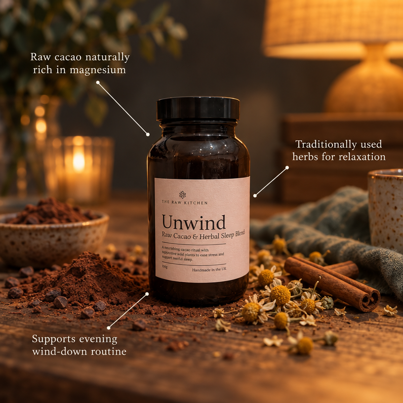 A dark amber bottle labeled 'Unwind' by 150 Raw Kitchen sits on a wooden surface with loose cacao powder, cinnamon sticks, dried flowers, and warm candlelight in the background. Text callouts describe the product benefits.