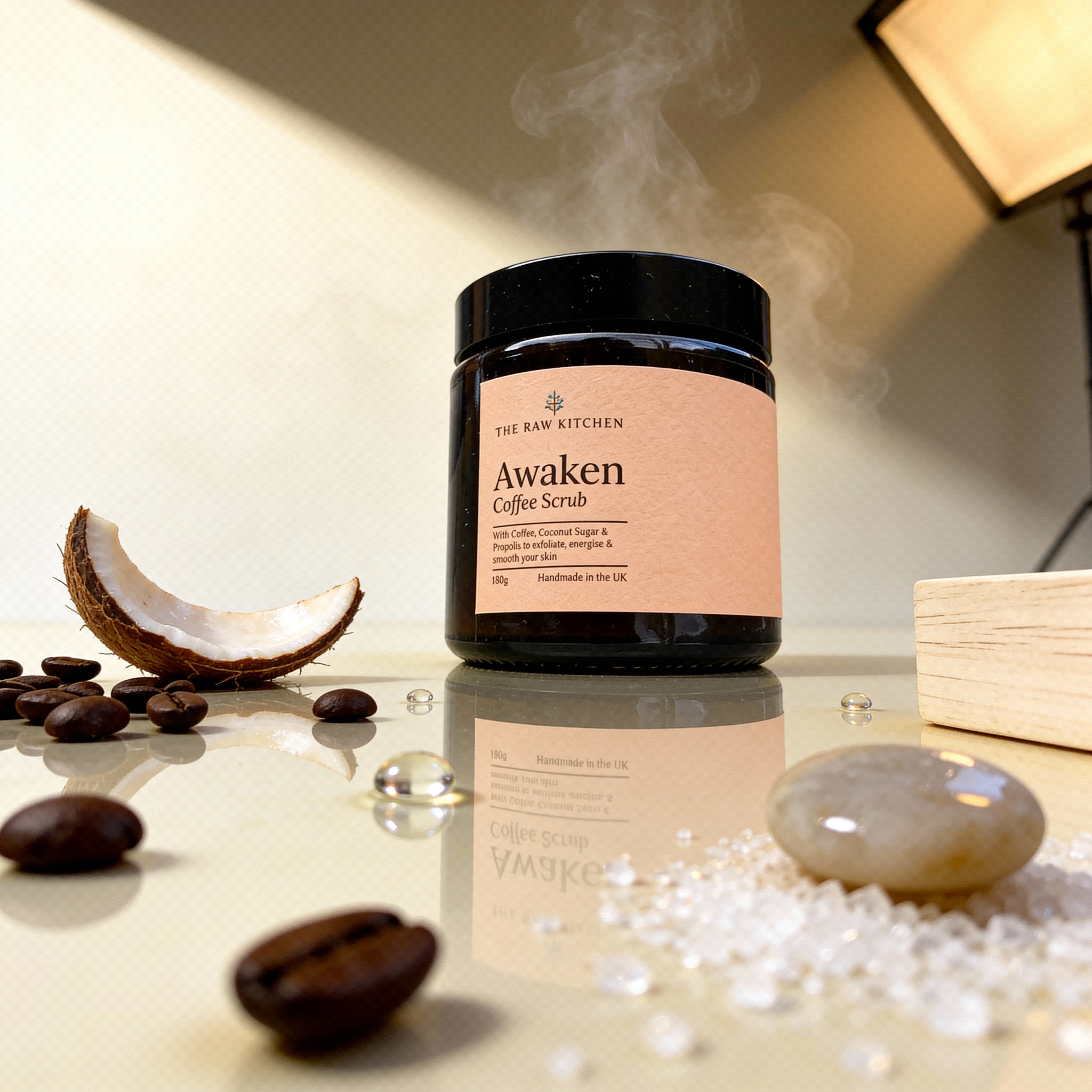 Jar of 'Awaken Coffee Scrub' from 'The Raw Kitchen' on a reflective surface with coffee beans and coconut pieces.
