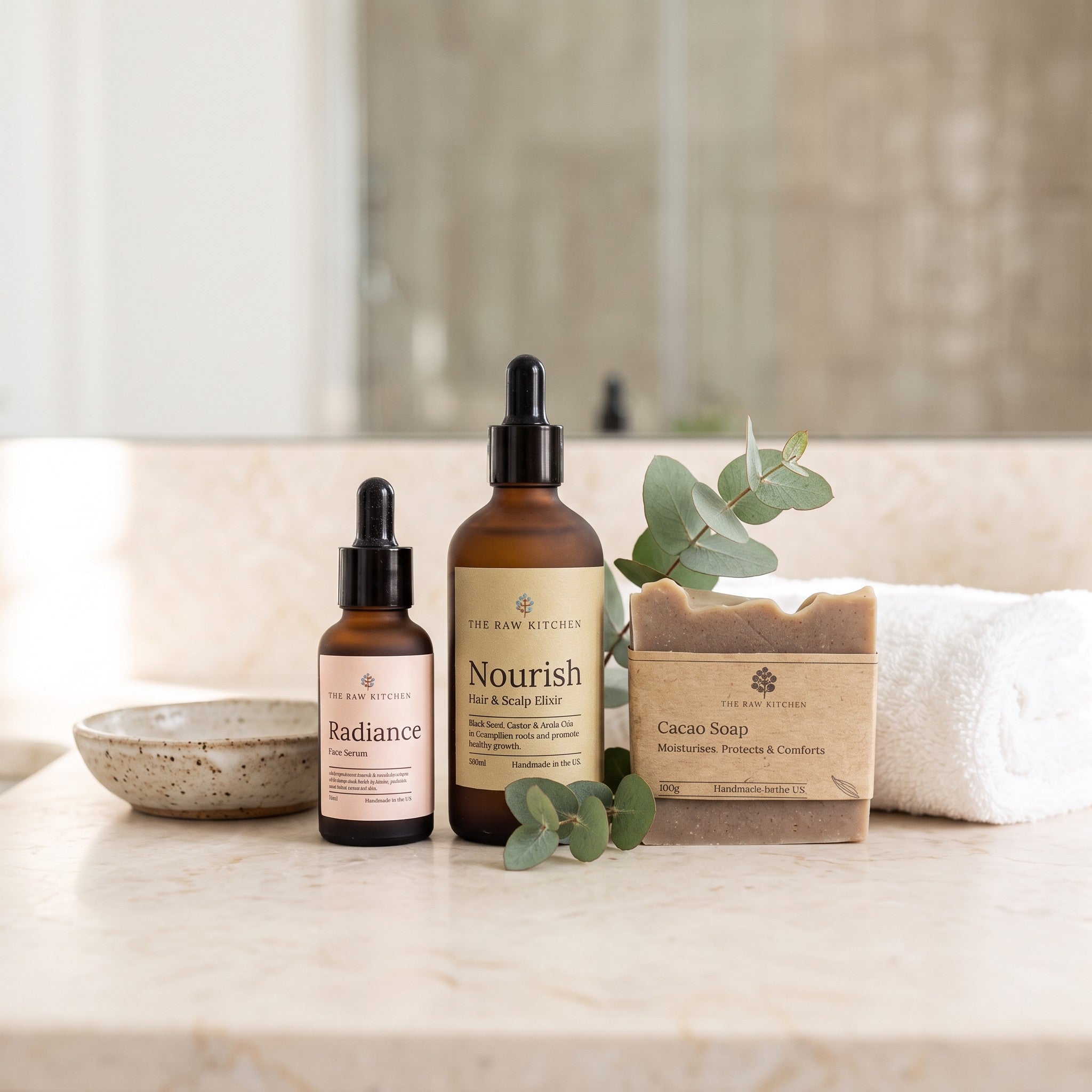 Three skincare products from The Raw Kitchens arranged on a marble countertop with a towel, bowl, and greenery in a softly lit bathroom setting: Radiance serum, Nourish face & body wash, and Cacao Soap.