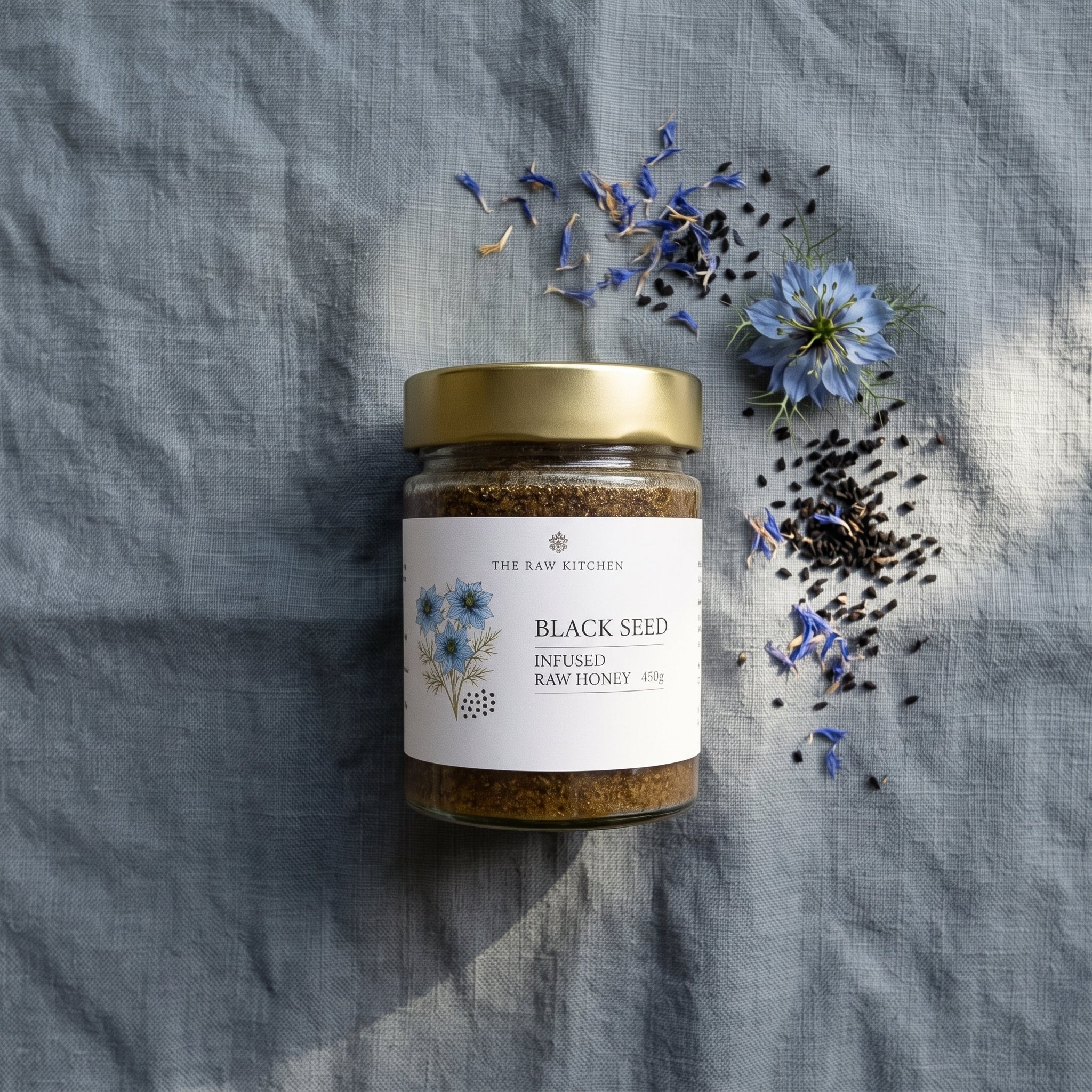 A glass jar of 'The Raw Kitchen' Black Seed Infused Raw Honey sits on a textured gray linen cloth, surrounded by scattered black seeds, blue flowers, and dried petals under soft natural light.